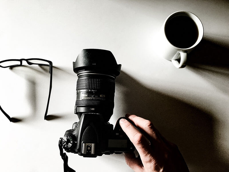 A man places a DSLR camera on a table next to a cup of black coffee and pair of reading glasses