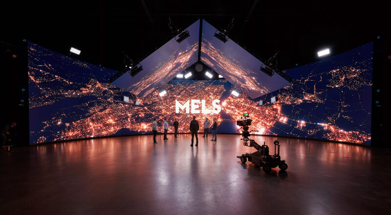 Le logo du studio MELs s'affiche au-dessus d'un paysage urbain sur une scène de production virtuelle.
